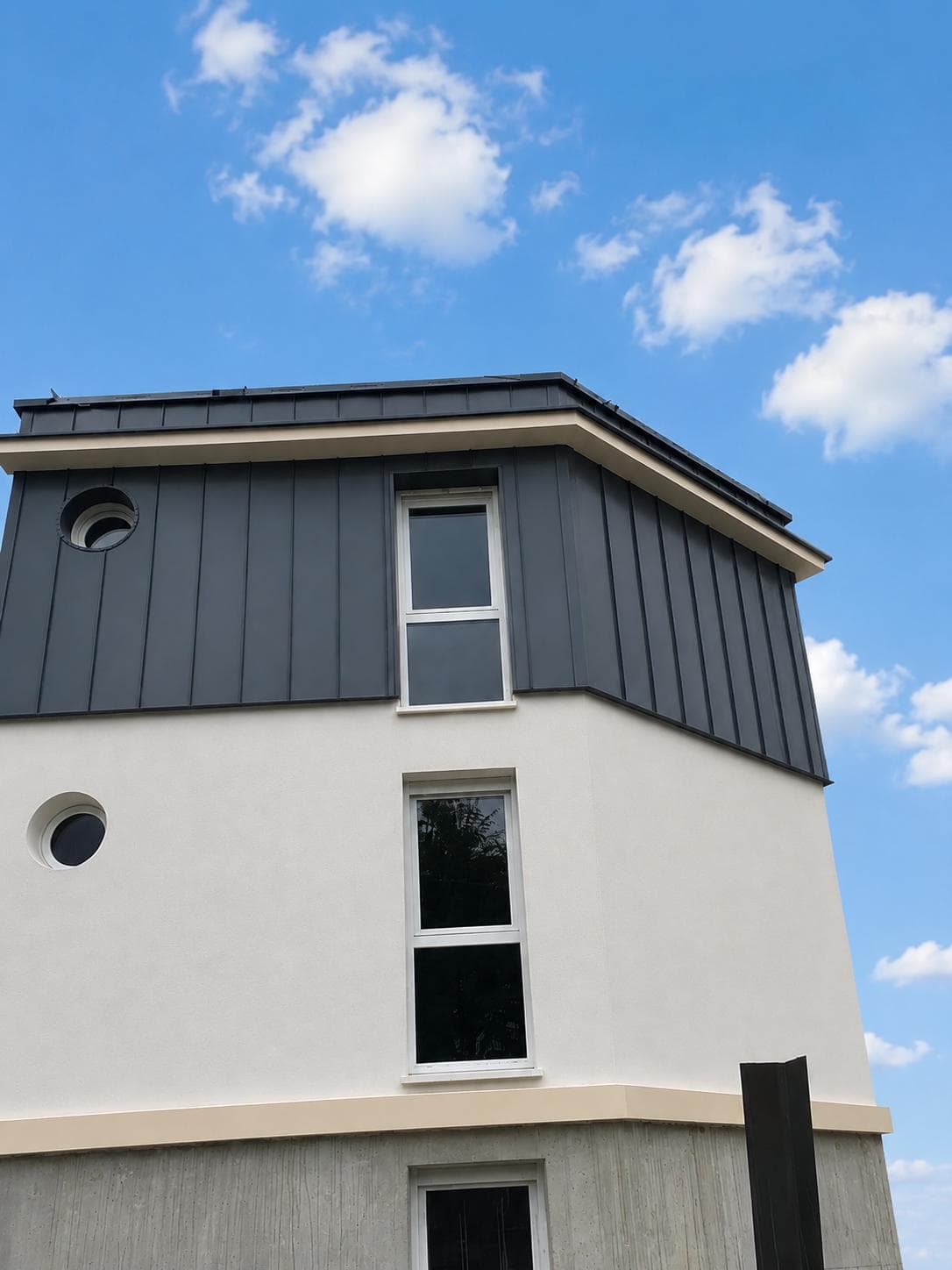 Bardage vertical en tôle anthracite habillant l'étage en surplomb d'un bâtiment clair.