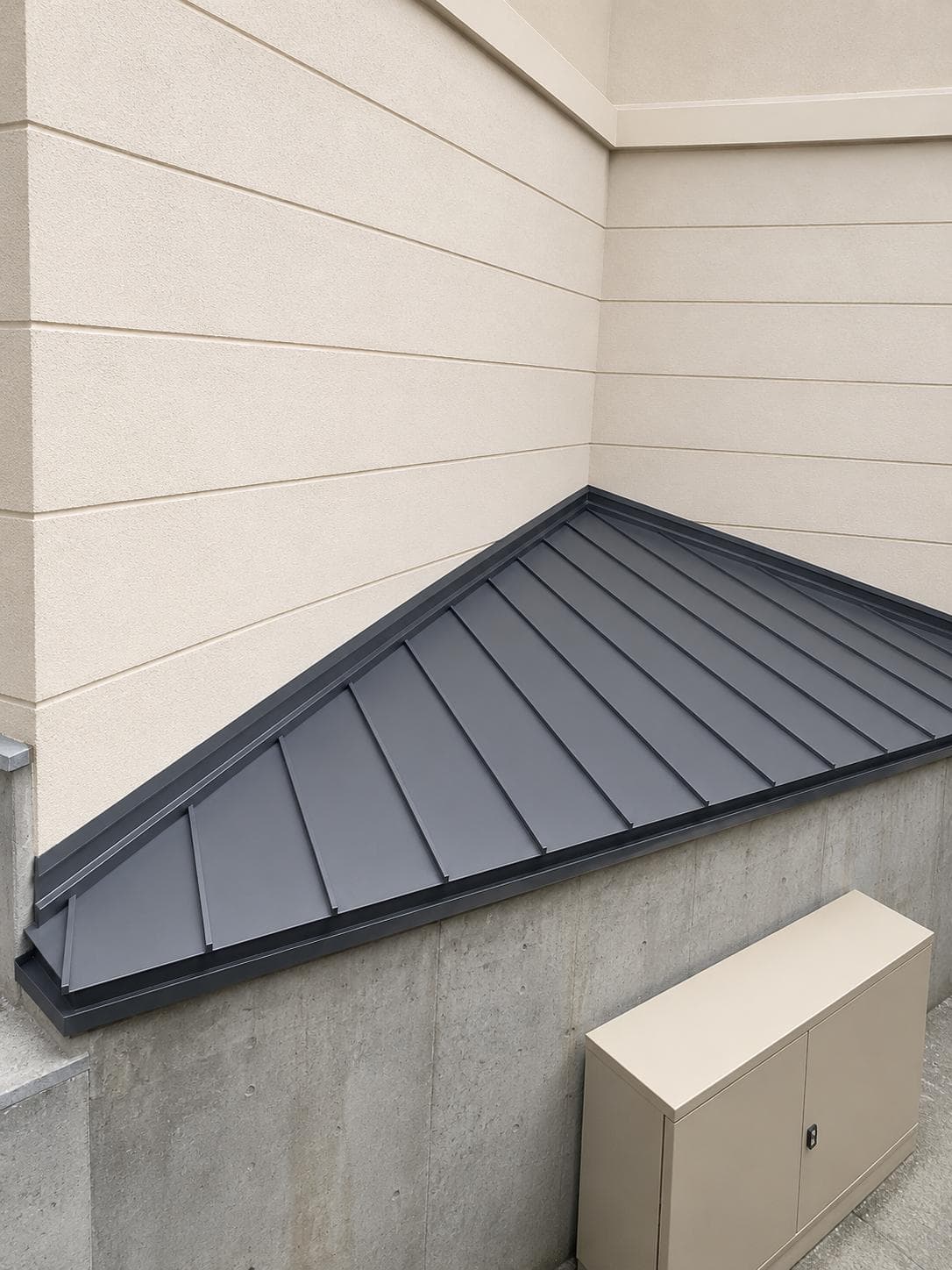 Petite couverture en tôle pliée anthracite à joint debout, en appui contre un mur de façade beige.