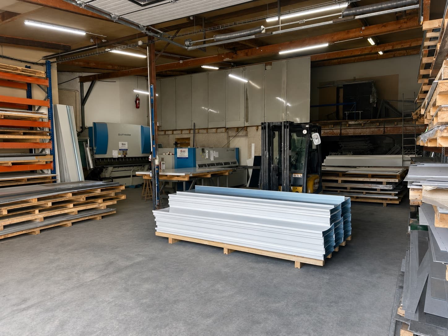 Pièces pliées en tôle laquée blanche, sous film de protection bleu, empilées sur palette devant les racks de tôles de l'atelier.